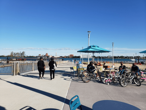 Two people walk along a waterfront promenade with tables, umbrellas, and bicycles nearby under a clear blue sky.