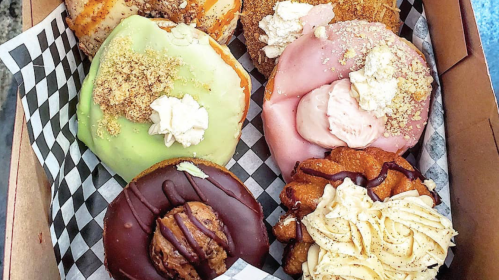 A box of six colorful donuts with various toppings, including frosting, whipped cream, and crumbs.