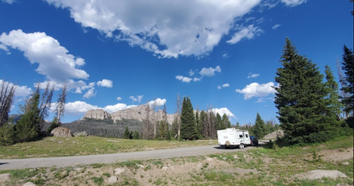 A scenic view of mountains under a blue sky with clouds, featuring an RV parked on a grassy area.