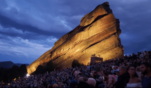 A large rock formation illuminated at dusk, with a crowd gathered in an outdoor amphitheater below.