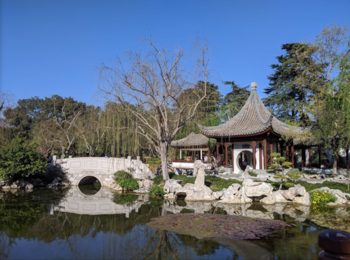 A serene garden scene featuring a traditional pavilion, a stone bridge, and lush greenery reflected in a pond.