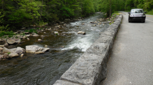 A winding river flows beside a stone wall, with a car parked on the roadside amidst lush green trees.