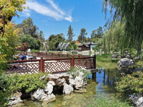 A serene garden scene featuring a pond, wooden bridge, lush greenery, and a traditional building under a clear blue sky.