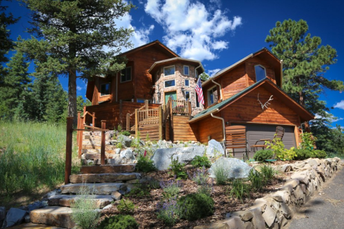 A rustic wooden house surrounded by greenery, featuring a stone pathway and a flag on the porch under a blue sky.