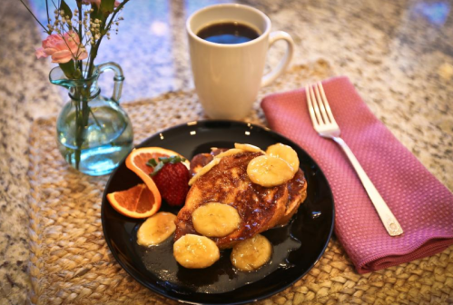 A plate of French toast topped with banana slices, served with strawberries, orange slices, and a cup of coffee.
