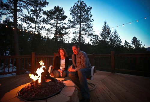 A couple sits by a fire pit on a deck, surrounded by trees, enjoying the evening glow and warm atmosphere.