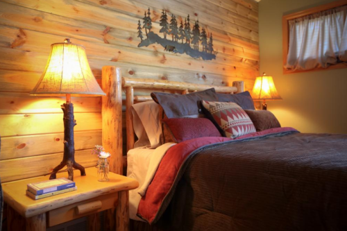 Cozy bedroom with a wooden bed, warm lighting, and rustic decor featuring a forest silhouette on the wall.
