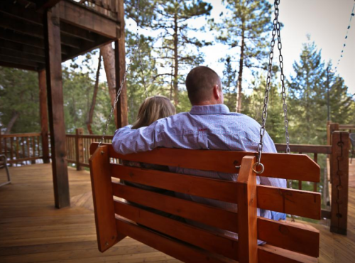 A couple sits on a wooden swing, embracing, surrounded by trees and a serene outdoor setting.