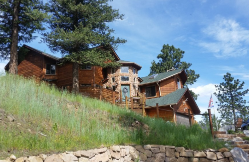 A wooden house on a hillside, surrounded by trees and grass, with a stone facade and a flag in the foreground.