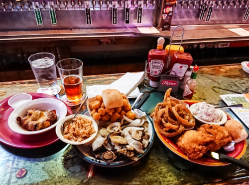 A table with various dishes: fried chicken, onion rings, tater tots, seafood, and drinks in a bar setting.
