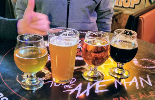 Four glasses of different beers on a table, with a person giving a thumbs up in the background.