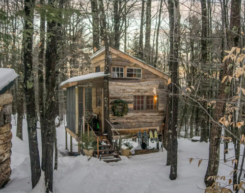 A cozy log cabin nestled in snowy woods, surrounded by trees, with a warm light glowing from the windows.