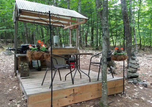 A rustic outdoor seating area with a wooden deck, table, chairs, and potted flowers, surrounded by trees.