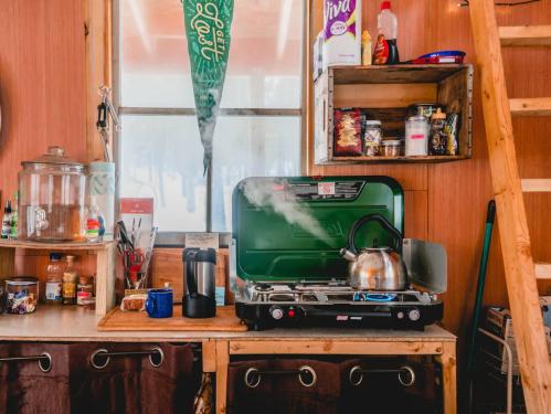 A cozy kitchen with a green camping stove, kettle steaming, and shelves filled with jars and food items.