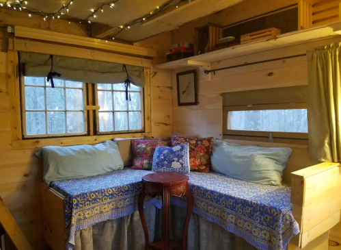Cozy interior of a wooden cabin with a seating area, colorful pillows, and warm lighting from string lights.