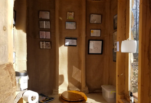 Interior of a small, rustic space with burlap walls, framed artwork, a wooden stool, and a container on the floor.
