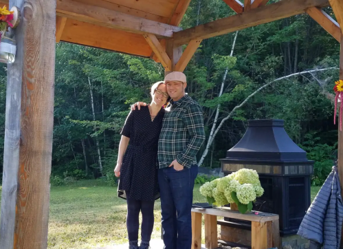 A couple stands together under a wooden gazebo, smiling, with greenery and a fireplace in the background.