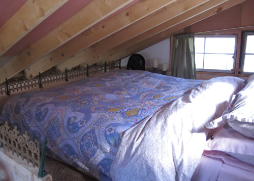 Cozy attic bedroom with a large bed covered in a patterned blanket and sloped wooden ceiling. Natural light from a window.