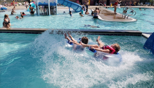 Two children slide into a pool on a float, splashing water as others swim nearby on a sunny day.