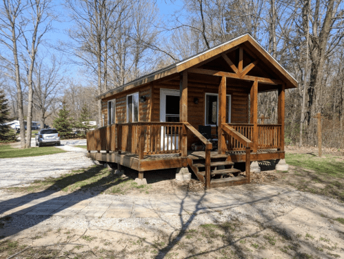 A small wooden cabin with a porch, surrounded by trees and a gravel path on a sunny day.