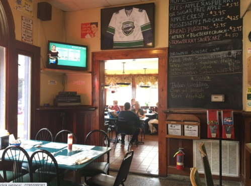 Cozy restaurant interior with tables, a chalkboard menu, and a TV displaying information. Diners are enjoying their meals.