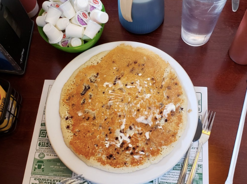 A large, golden-brown pancake with butter on a white plate, surrounded by condiments and a glass of water.