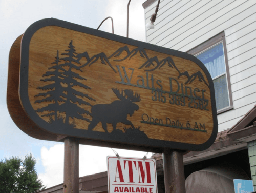 Sign for Walt's Diner featuring a moose and mountains, stating "Open Daily 6 AM" and an ATM available.