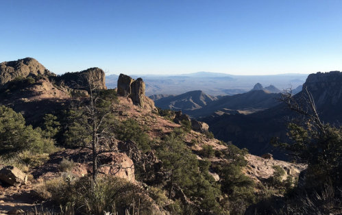 A panoramic view of rugged mountains and valleys under a clear blue sky, with sparse vegetation in the foreground.