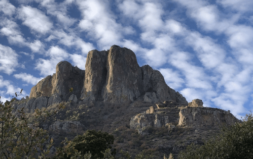 A rugged mountain peak rises against a blue sky with scattered clouds, surrounded by greenery and rocky terrain.
