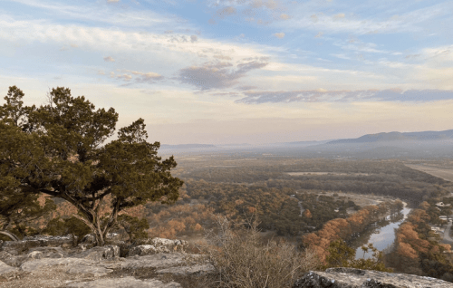 A scenic view from a rocky overlook, featuring rolling hills, a winding river, and autumn-colored trees under a cloudy sky.