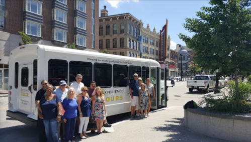A group of people poses next to a tour bus on a city street, with historic buildings in the background.