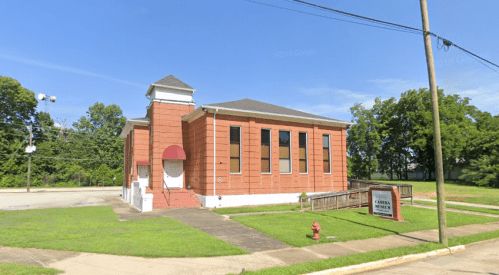 A brick building with large windows and a red entrance, surrounded by green grass and trees.