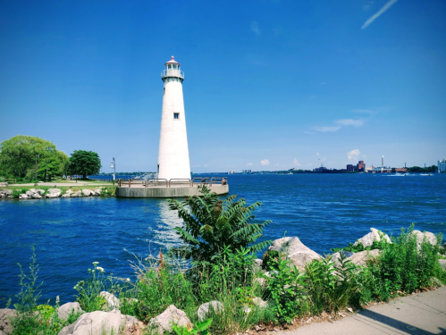 A tall white lighthouse stands by a blue lake, surrounded by greenery and rocky shoreline under a clear blue sky.