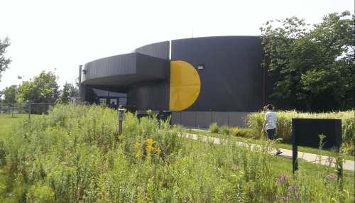 A modern building with a large yellow circle on its facade, surrounded by greenery and a walking path.