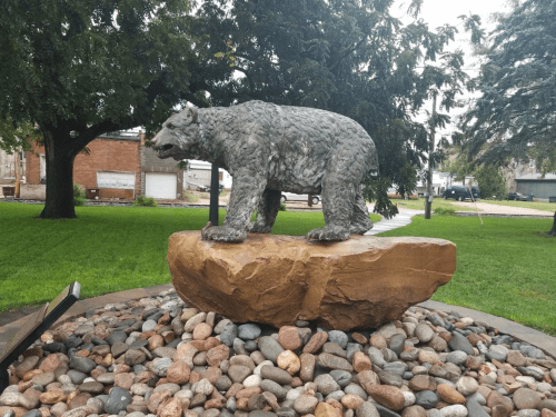 A bear statue stands on a large rock, surrounded by colorful pebbles, in a grassy park setting.