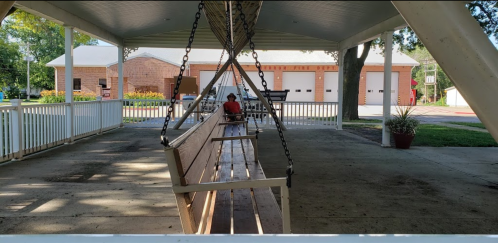 A person sits on a swing in a covered pavilion, with a fire station visible in the background.