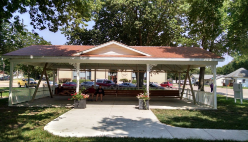 A covered pavilion with benches, surrounded by trees and flowers, in a park setting with cars in the background.