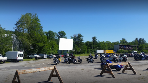 A parking lot filled with motorcycles, a large blank screen, and trees in the background on a sunny day.