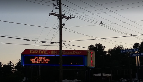A drive-in theater sign displays "24 Hour Movie Line" with a phone number, set against a twilight sky.