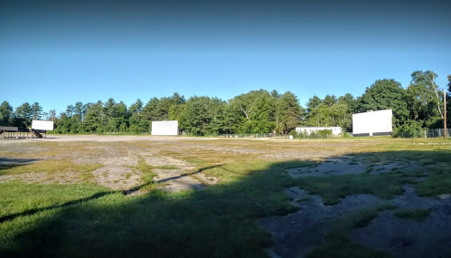 Abandoned drive-in theater with empty screens and overgrown grass, surrounded by trees under a clear blue sky.