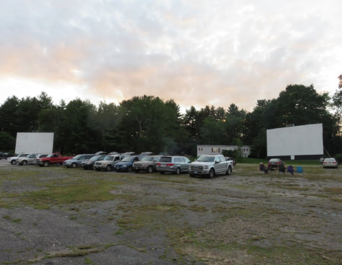 A drive-in movie theater with parked cars and two large screens, surrounded by trees under a cloudy sky.