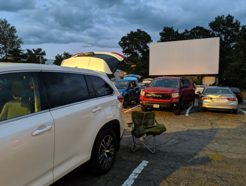 A view of a drive-in movie theater parking lot with cars and a large screen in the background under a cloudy sky.