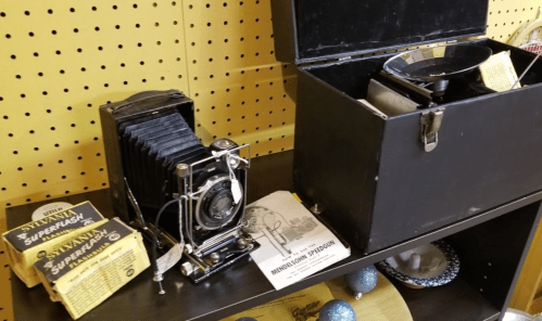 A vintage camera with film boxes and a black storage case on a shelf, alongside a brochure for a microscope.
