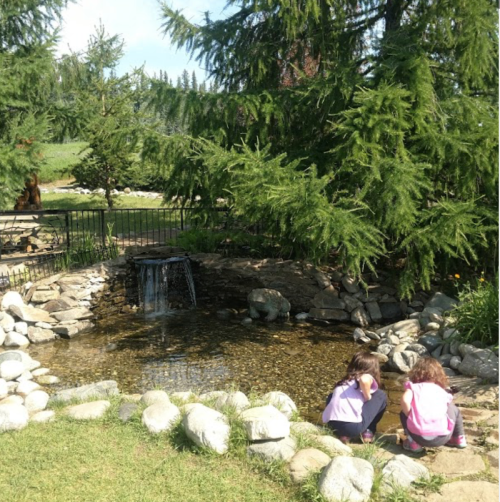 Two children sit by a small pond surrounded by rocks and trees, playing near a gentle waterfall.