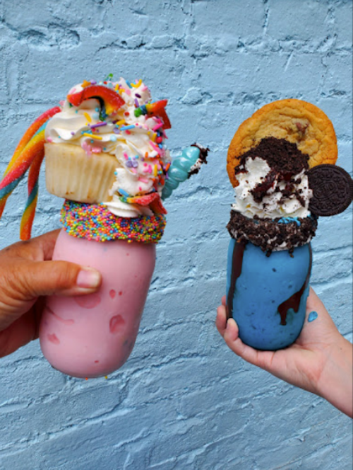 Two colorful milkshakes in jars, one pink topped with a cupcake and sprinkles, the other blue with cookies and whipped cream.