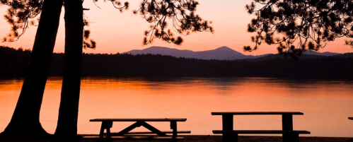 Silhouette of trees and picnic tables by a calm lake at sunset, with mountains in the background and a colorful sky.