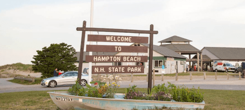 Welcome sign for Hampton Beach State Park, with a boat planter and a building in the background.