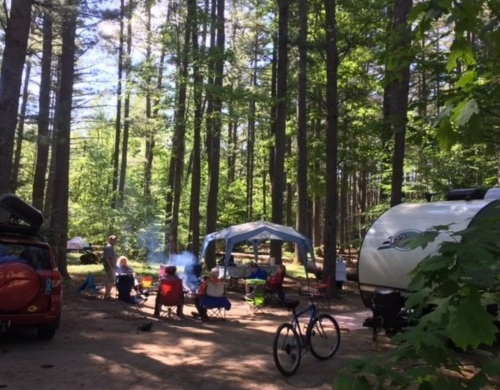 A campsite in a forest with people gathered around a fire, tents, and a bicycle nearby.