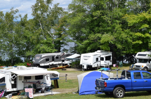 A scenic campsite by a lake with RVs, tents, and kayaks surrounded by trees under a partly cloudy sky.