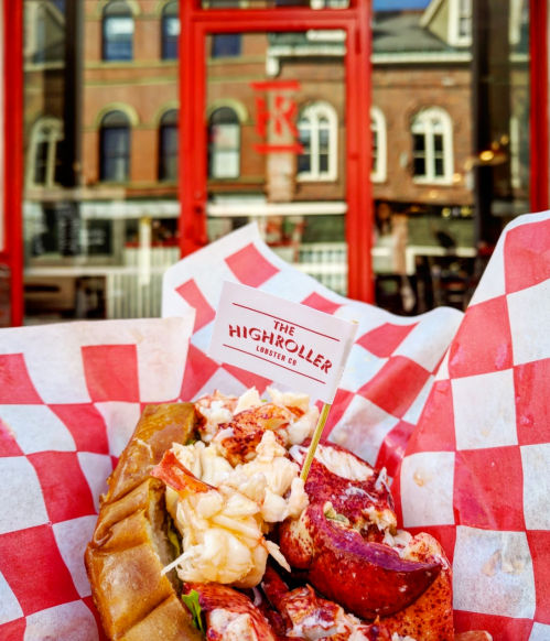 A lobster roll with a flag reading "The Highroller Lobster Co." in front of a red storefront.